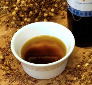 High Lignan flaxseed Linseed oil with dark particles in an amber coloured oil contained in a ramekin next to a bottle and flaxseeds.